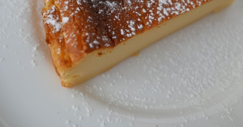 A wedge of pale baked custard flan with a browned, sugar-dusted top on a white plate