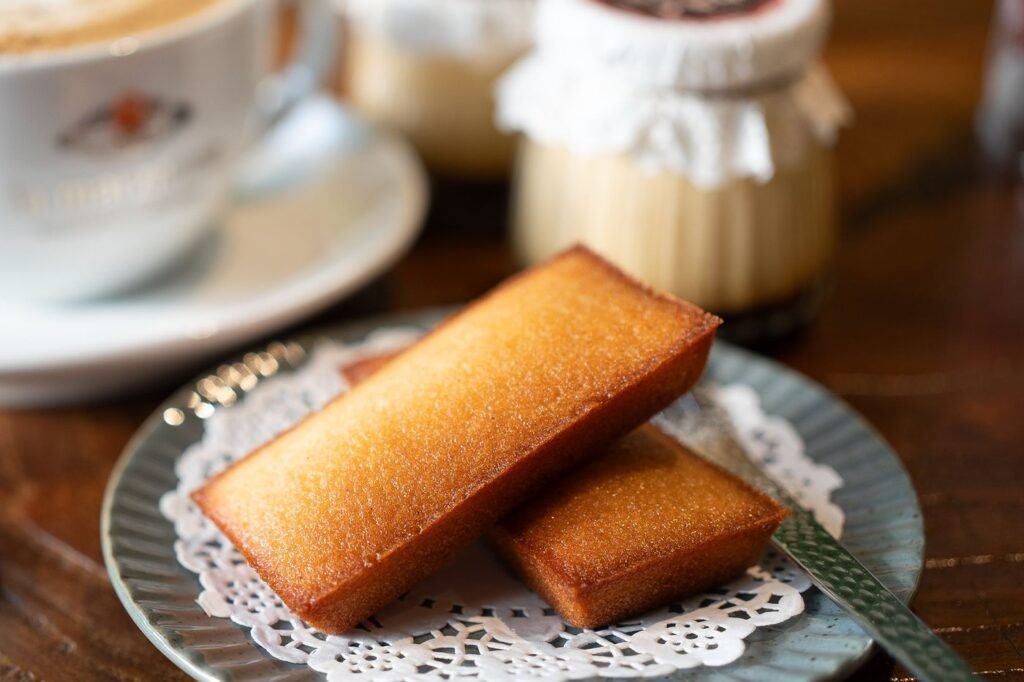 Two golden financiers, the flat rectangular French almond cakes, on a paper doily with a cup of coffee behind